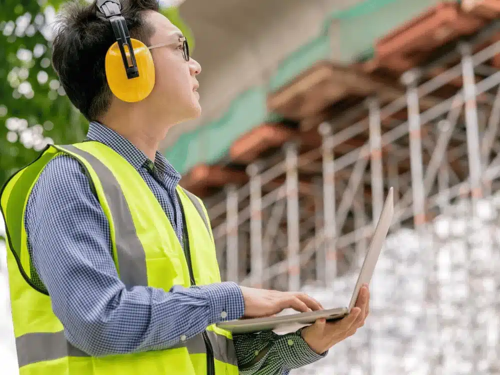 Construction worker wearing safety gear and ear protection, using a laptop at a construction site.
