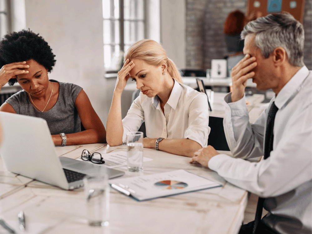 A group of business people sitting at a table with their heads in their hands.