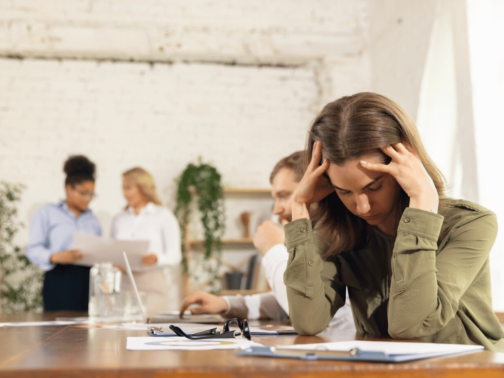 A person sitting at a table with their head in their hands and people in the background.