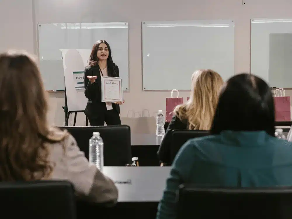 A presenter holds a certificate while speaking to an engaged audience in a conference room, with whiteboards in the background.