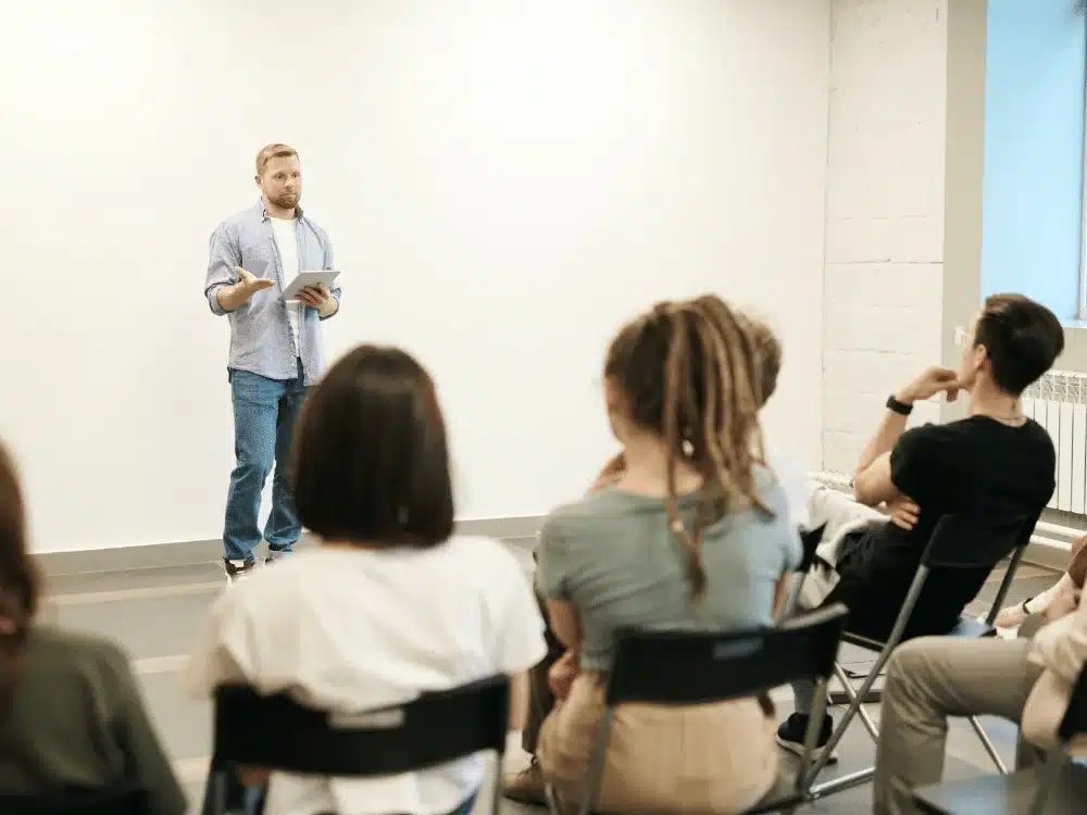 Business coaching event with a casual-dressed presenter speaking to seated adults.