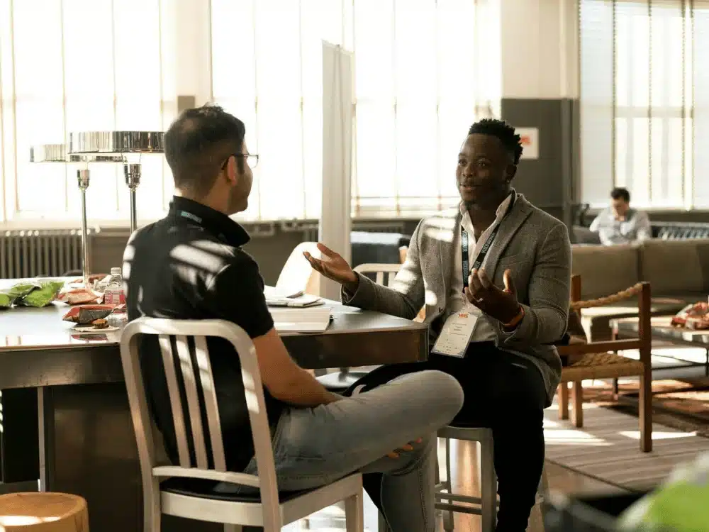 Two professionals engaged in a conversation at a conference table in a well-lit room.