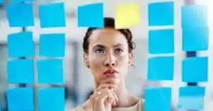 A person ponders in front of a wall covered in colorful sticky notes, with a focus on a bright yellow note among the blue ones.