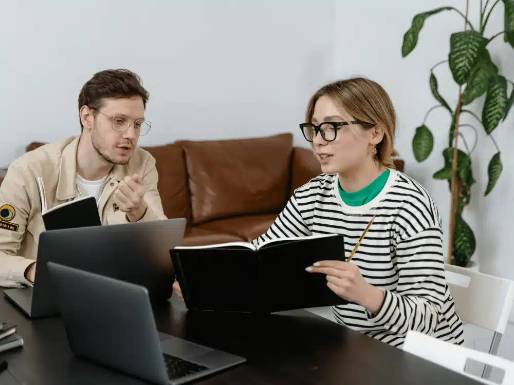 Two colleagues discussing a project while reviewing notes and working on laptops in a casual office setting.