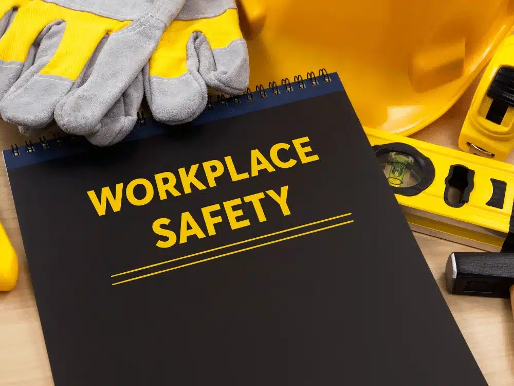 A black notepad titled "WORKPLACE SAFETY" surrounded by yellow tools and gray safety gloves, symbolizing construction safety.