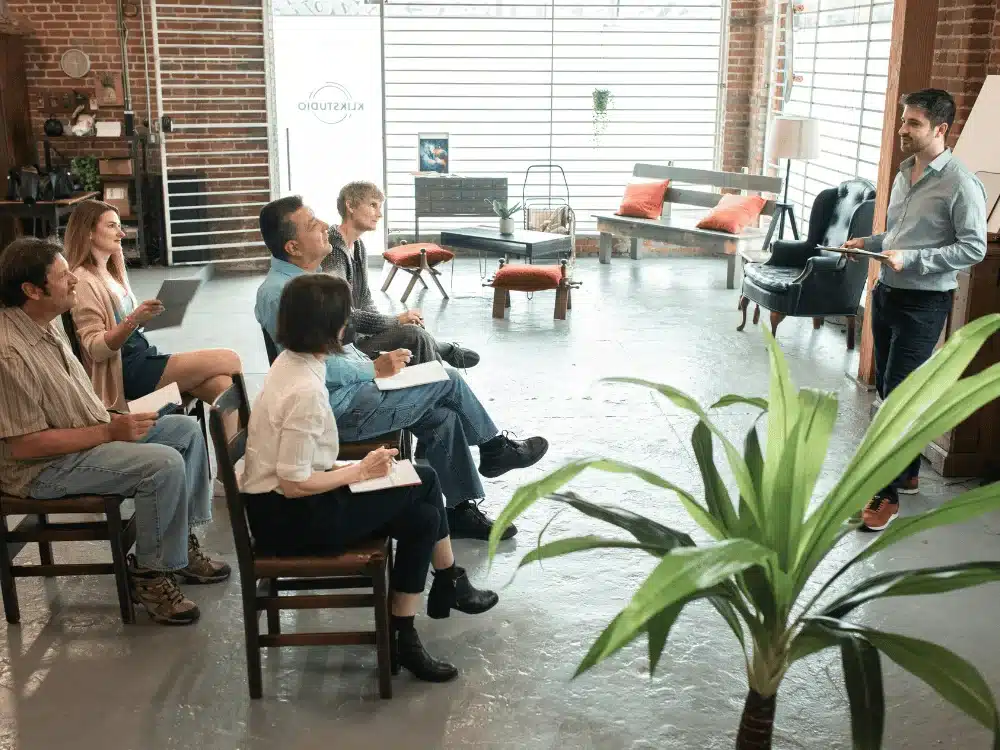 A group of six people attentively listens to a speaker in a bright, modern studio with plants and colorful furniture.