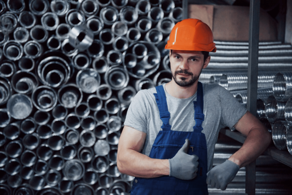 A person in an orange hard hat and overalls standing in front of stacks of pipes.
