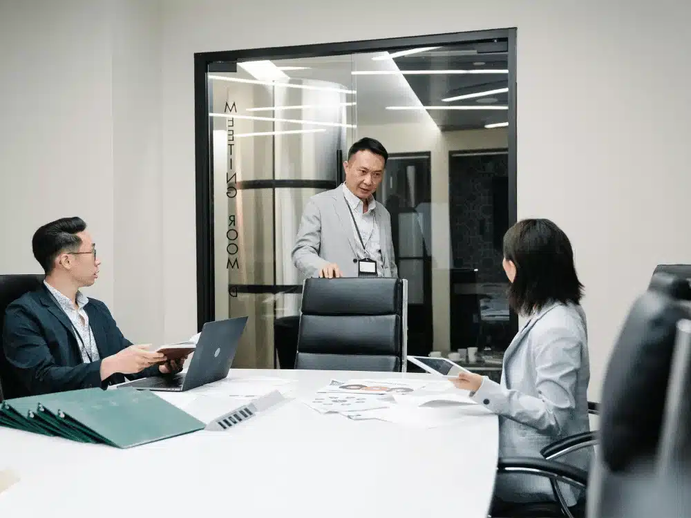 Colleagues in a conference room, with one person standing and two sitting with laptops and documents.