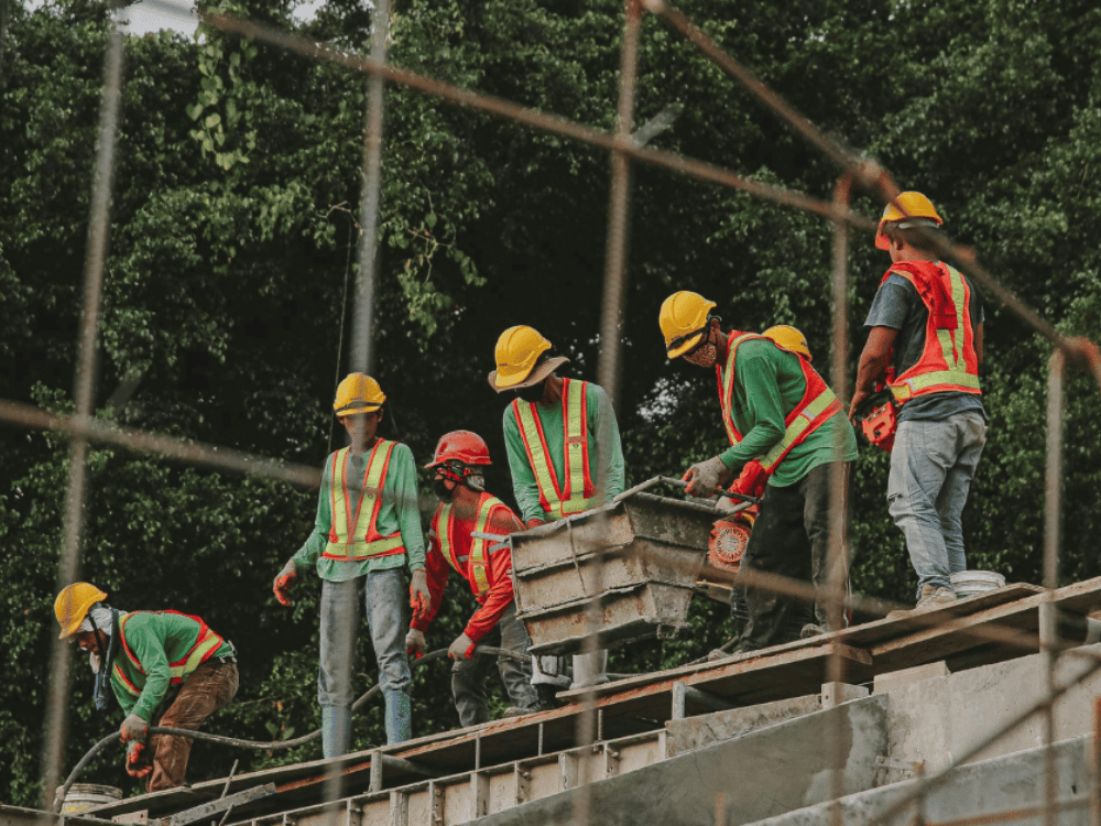 Construction workers in helmets and high-visibility vests focus on building a wall at a greenery-flanked site.