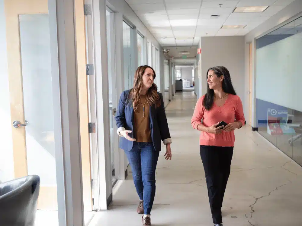 A candid moment between two female employees sharing ideas during a walk in a well-lit office hallway.
