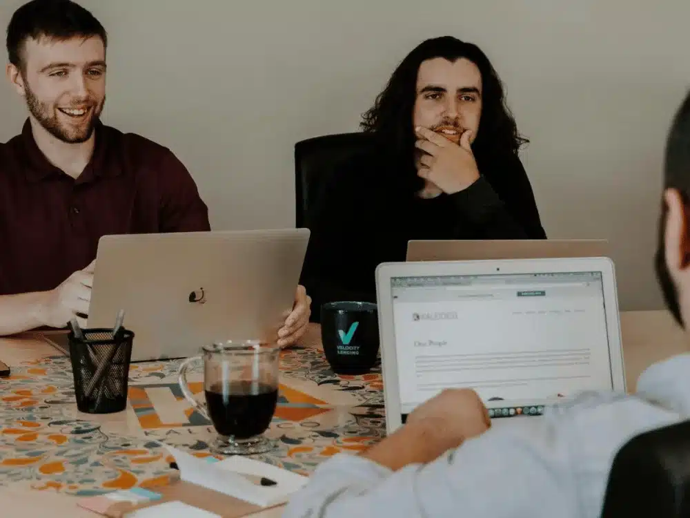 Three colleagues in a meeting, two with laptops, engaged in discussion.