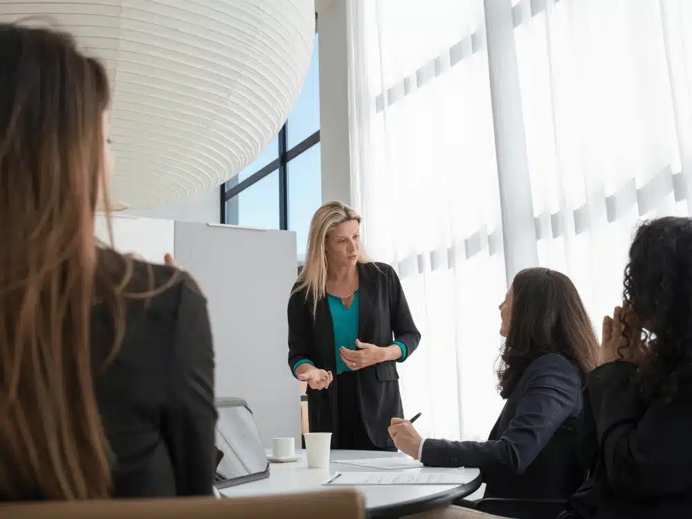 A woman presents to an engaged audience in a bright, modern conference room with large windows and stylish decor.