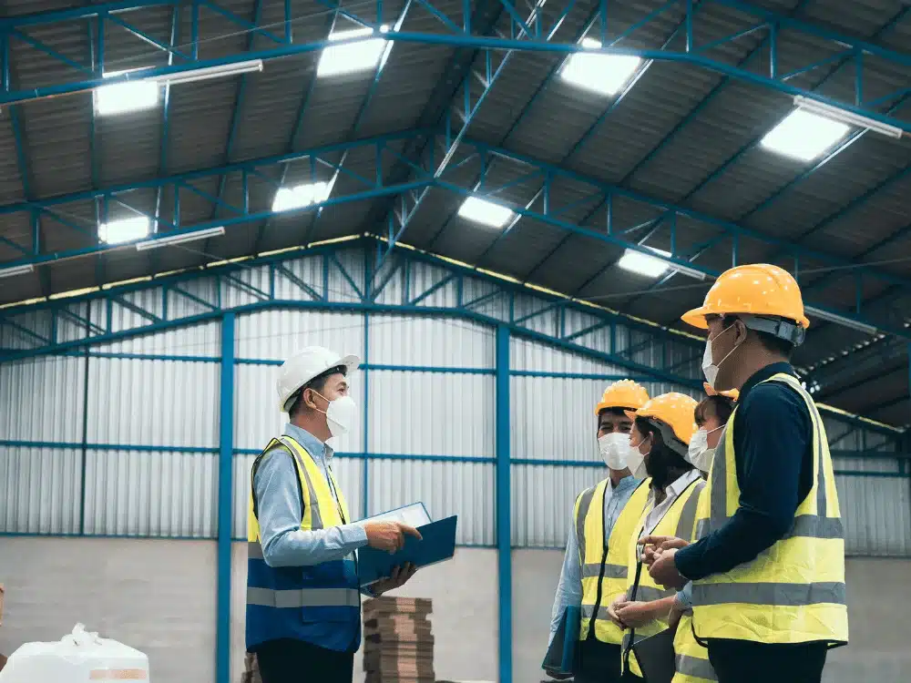 A group of workers in hard hats and safety vests, attending a safety briefing in a spacious warehouse with bright lighting.