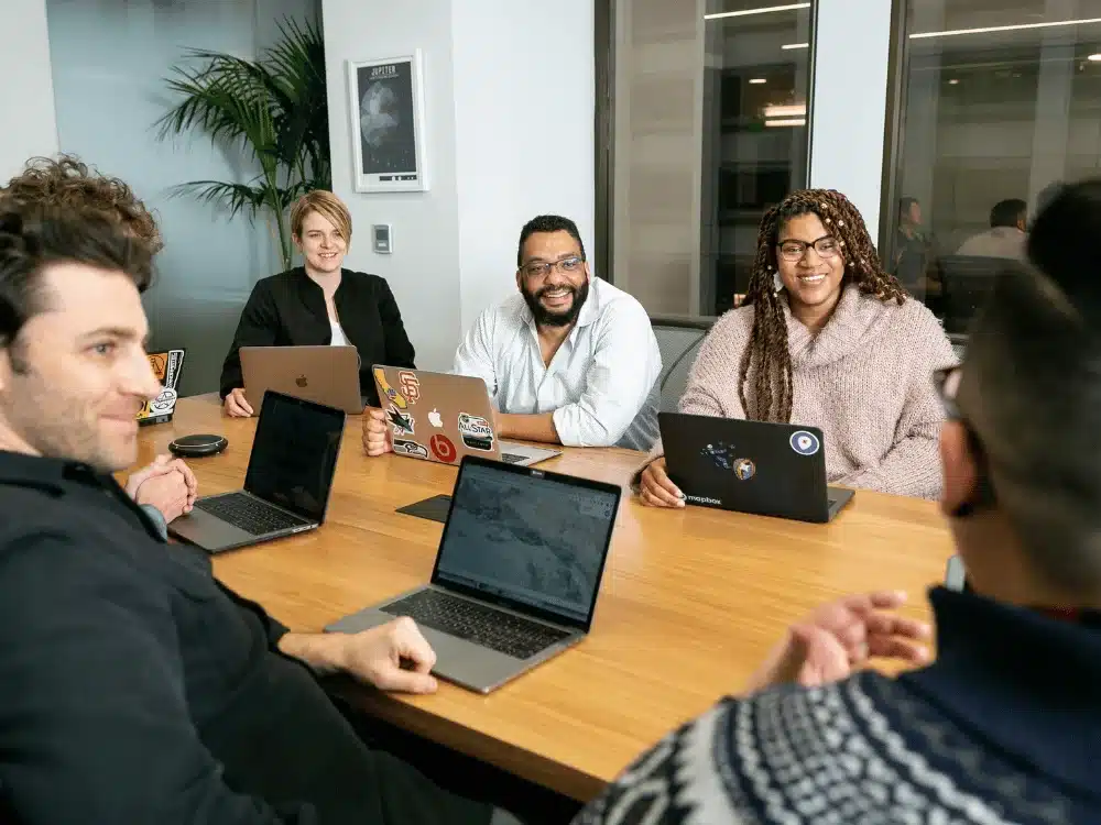 A group of professionals in a modern conference room, collaborating with laptops on a wooden table and greenery in the background.