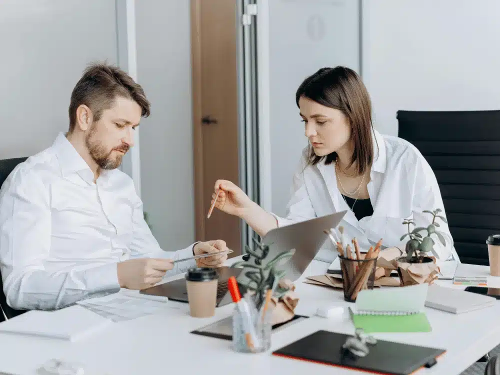 Two colleagues reviewing documents and working on a laptop in a modern office.