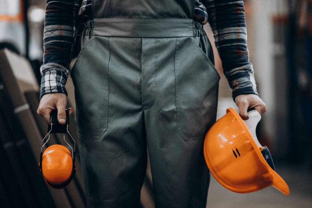A person in overalls holding an orange hard hat.