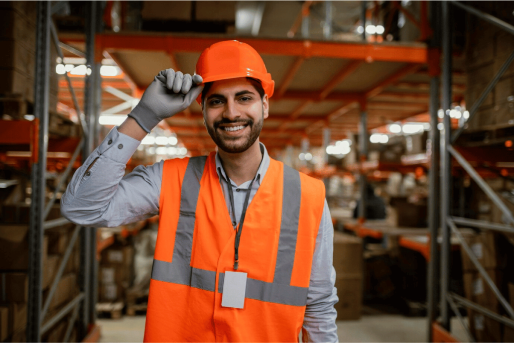 A person in an orange vest and hard hat smiling inside a warehouse.