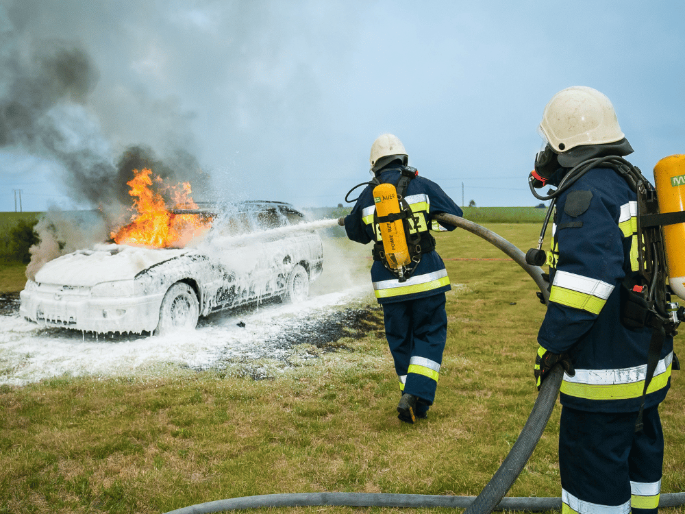 Firefighters in protective gear extinguishing a car fire in an open field.