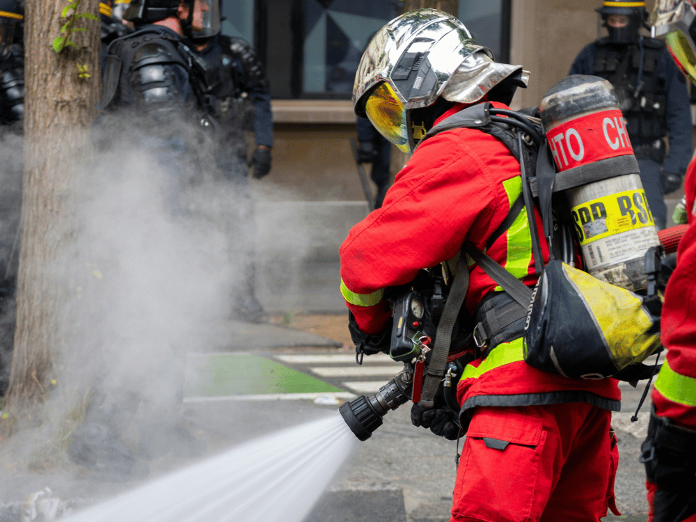 Professional firefighter aiming water hose at fire amidst city buildings.