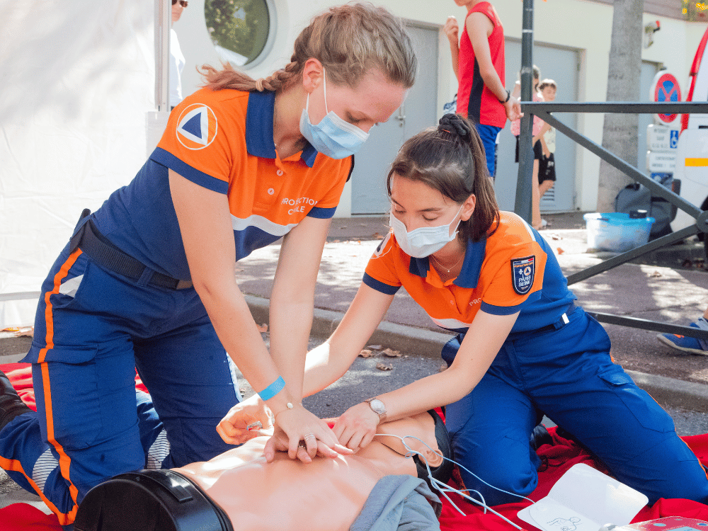 Paramedics in orange and blue uniforms practicing CPR on a mannequin outdoors.