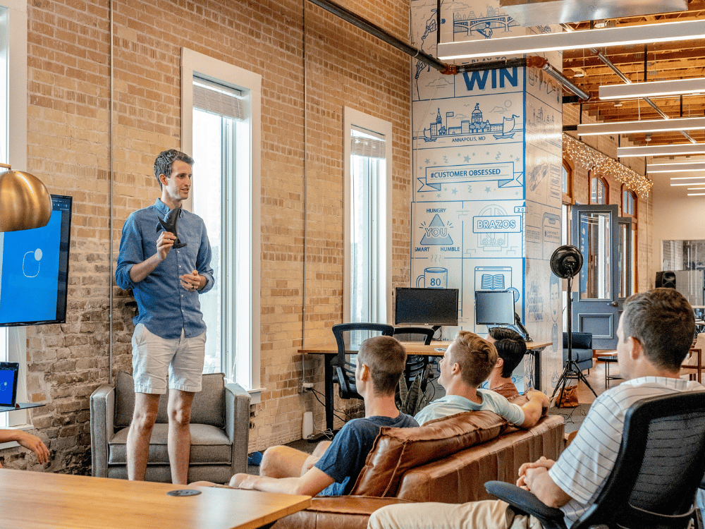 A man in a blue shirt and shorts presents at a company meeting, with attendees seated and attentive.
