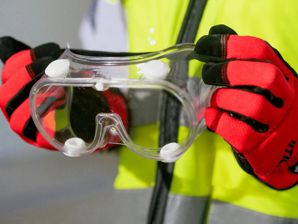 Close-up of safety goggles held by a worker with red and black gloves.