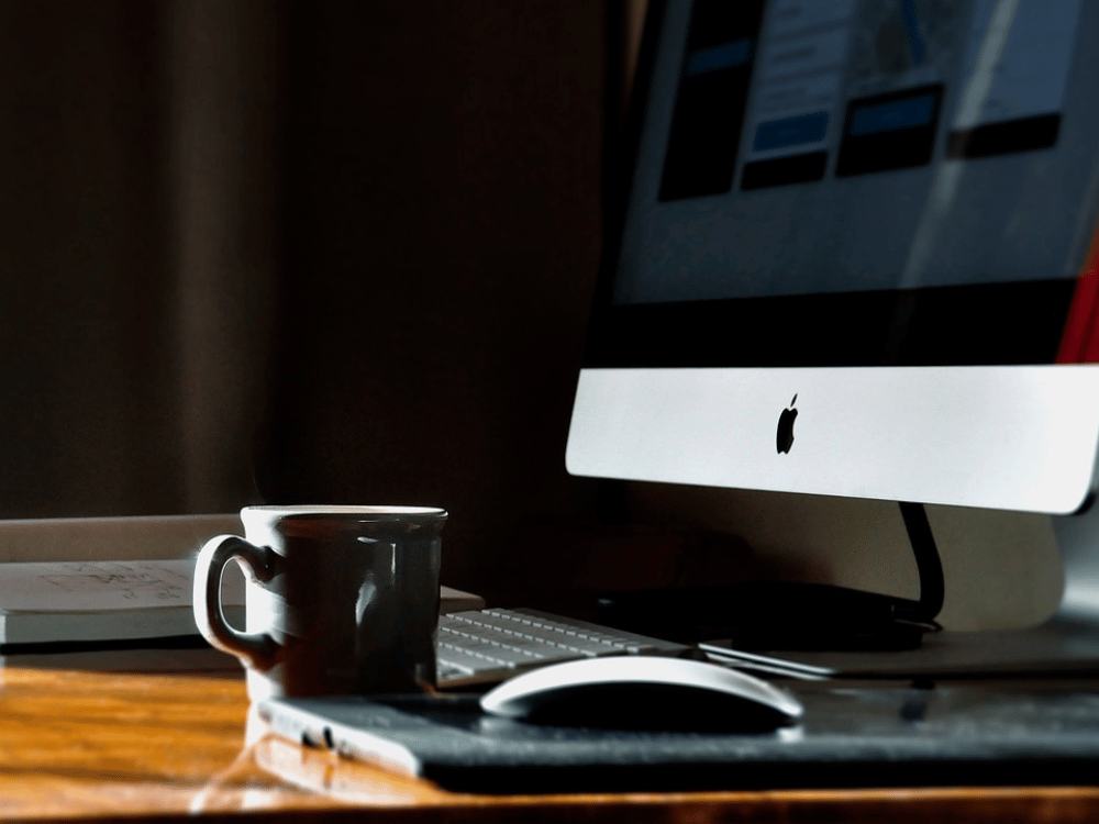An apple computer sits on a desk next to a cup of coffee.