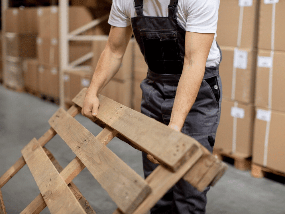 A person in overalls is holding a pallet in a warehouse.