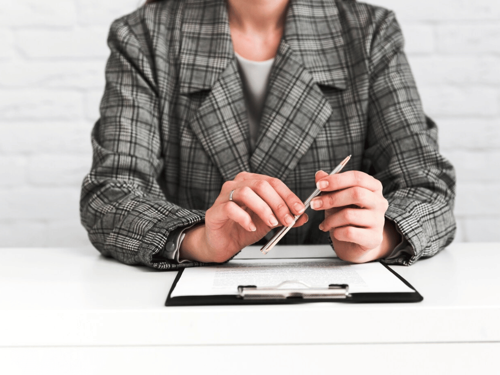 Professional in plaid blazer holding pen over clipboard on desk.