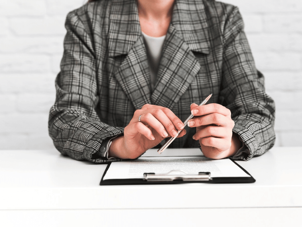 Professional in plaid blazer holding pen over clipboard on desk.
