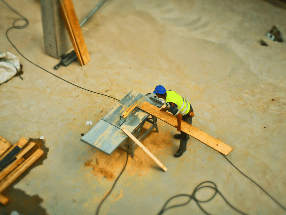 A construction worker wearing a high-visibility vest and hard hat while cutting wood on a workbench.