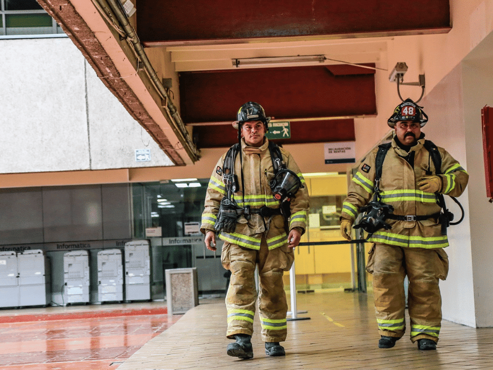 Firemen in protective suits carrying equipment indoors.