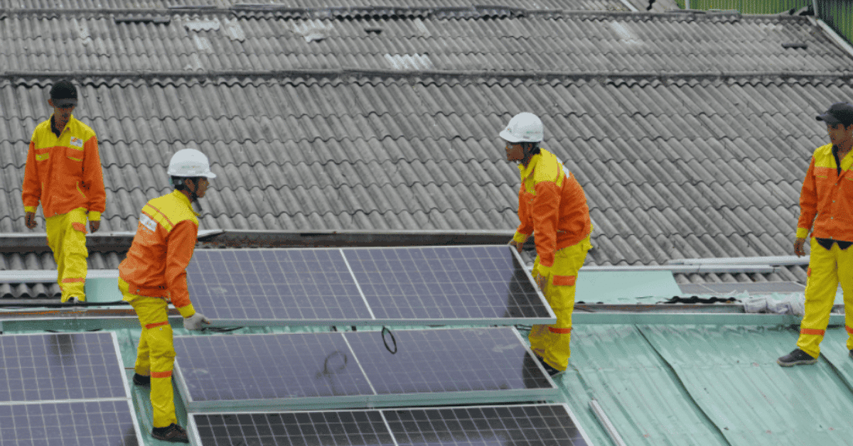 Four technicians in safety helmets and reflective jackets fitting photovoltaic modules on a rooftop.