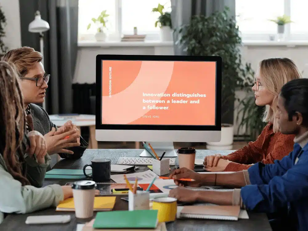 Team meeting around a table with a computer screen displaying a Steve Jobs quote.