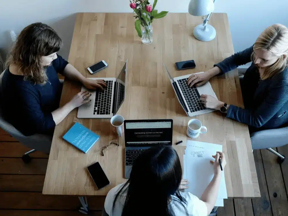 Three women working on laptops at a wooden table, viewed from above.