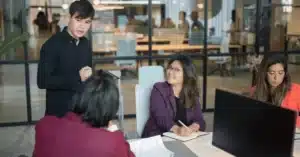 Four colleagues in a modern office engaged in a discussion, with one person standing and speaking.