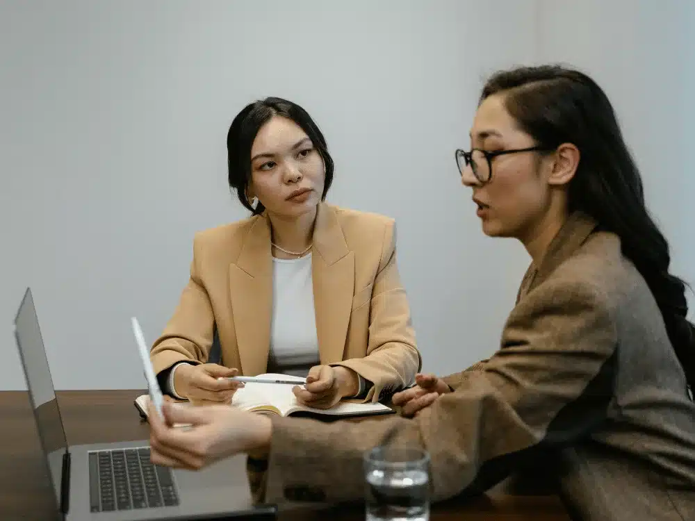 Two women in business attire having a serious discussion at a table with a laptop.