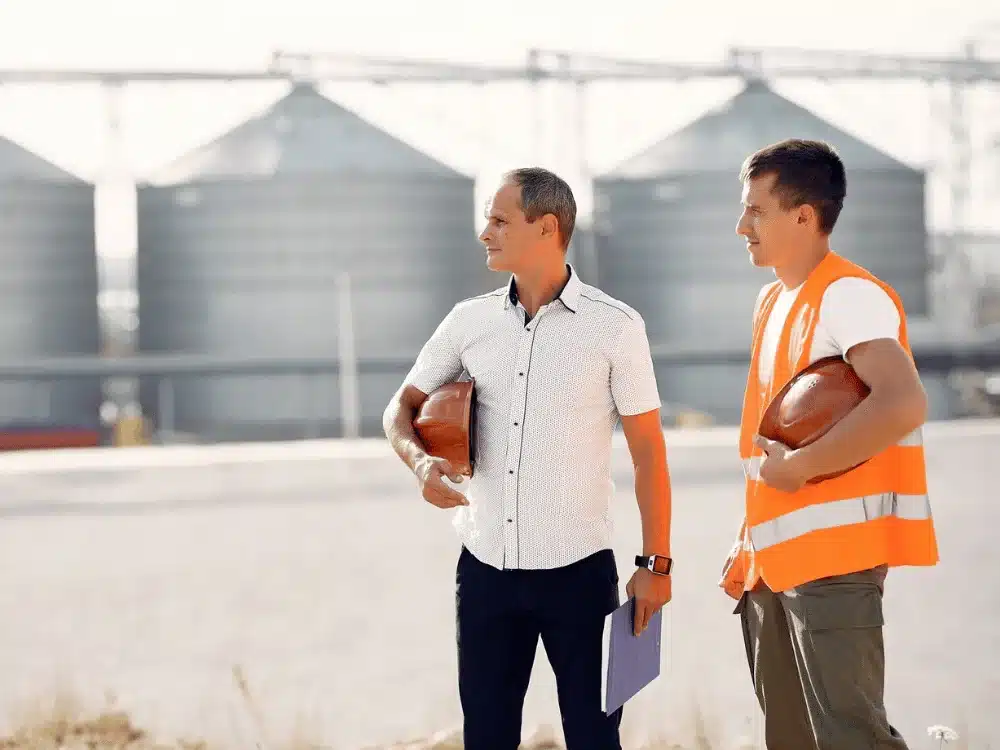 Two industrial workers with safety helmets discussing near large storage silos.