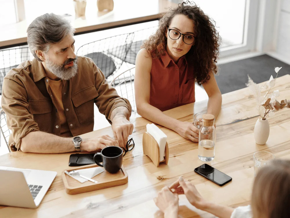 Three colleagues having a serious discussion at a wooden table in a bright office.