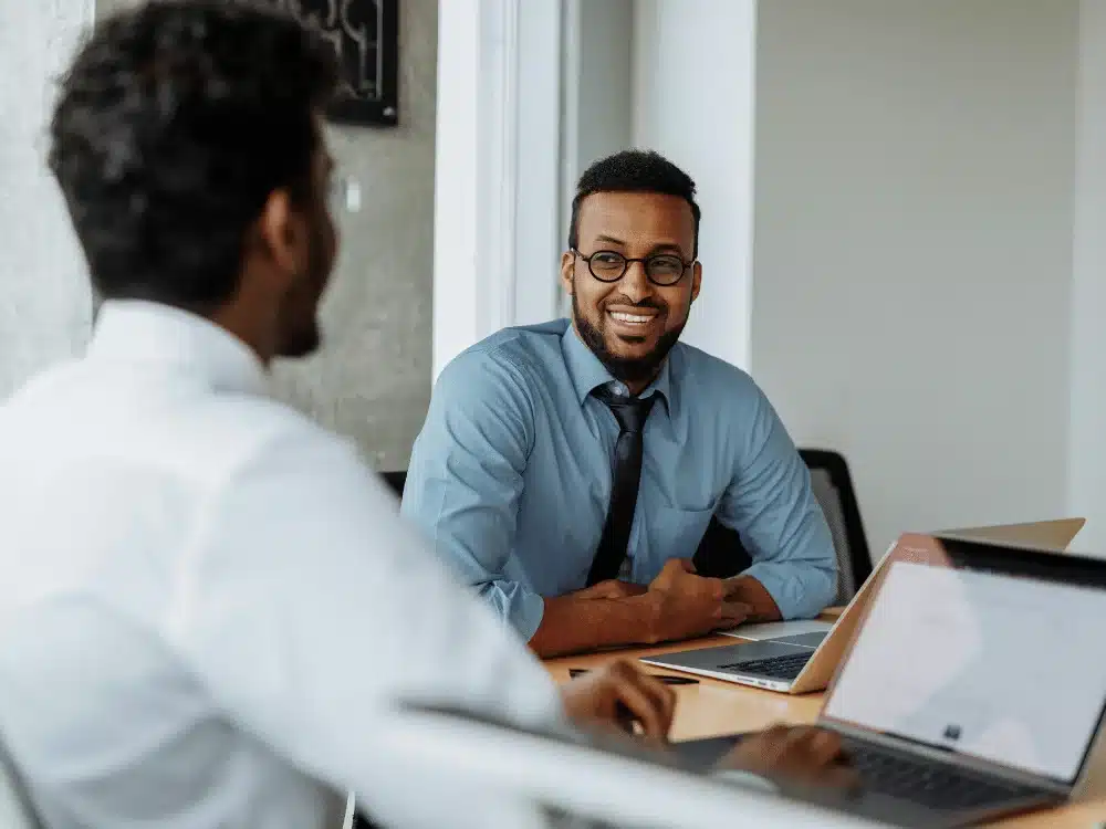 Smiling man in a blue shirt conversing with a colleague at a desk.