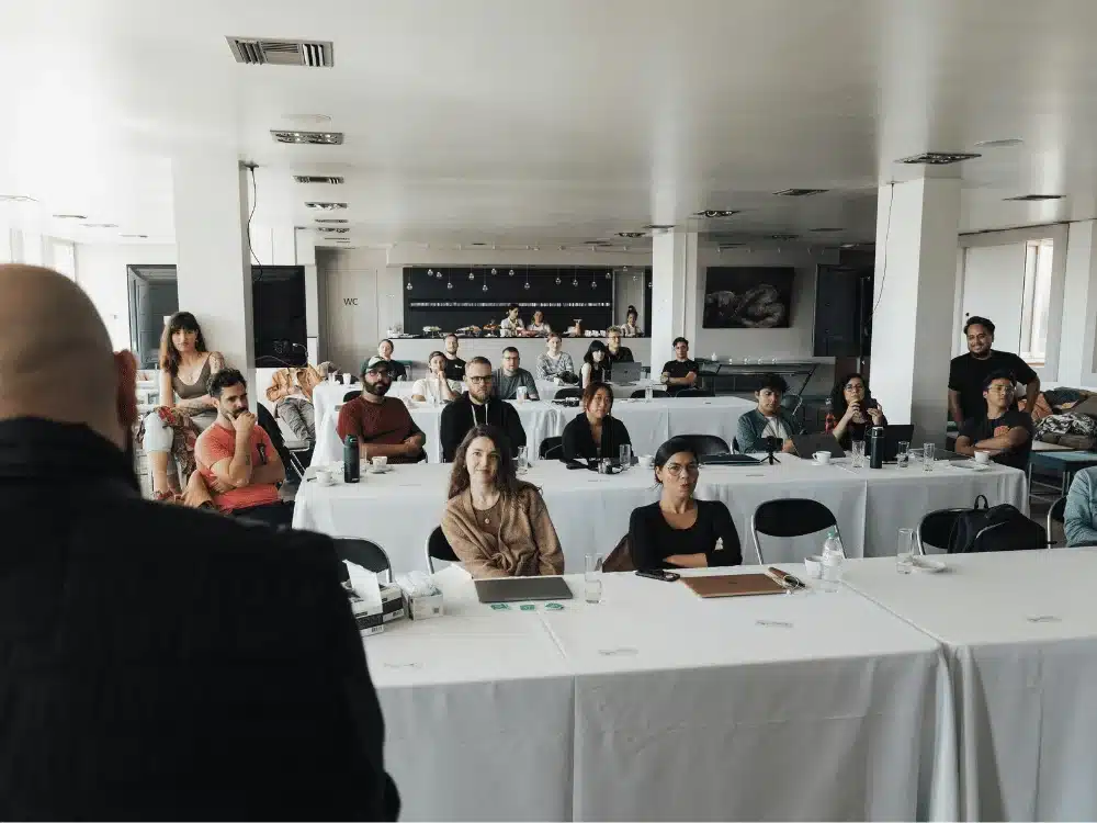 Audience attentively listening to a speaker during a conference in a large room with white tables.