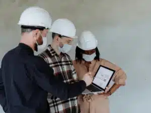 Engineers wearing hard hats and masks reviewing blueprints on a laptop.