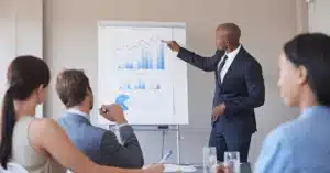 A professional man in a suit presents data on a graph to three attentive colleagues in a modern meeting room.