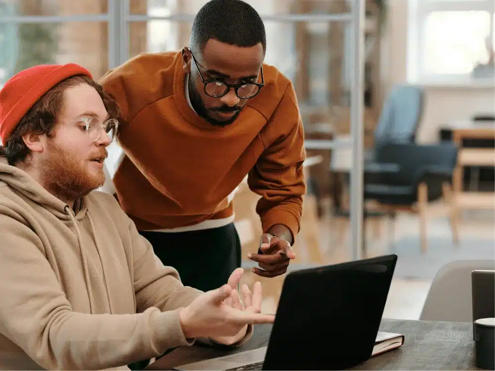 Two individuals engage in conversation while looking at a laptop, one gesturing as the other listens attentively in a modern workspace.