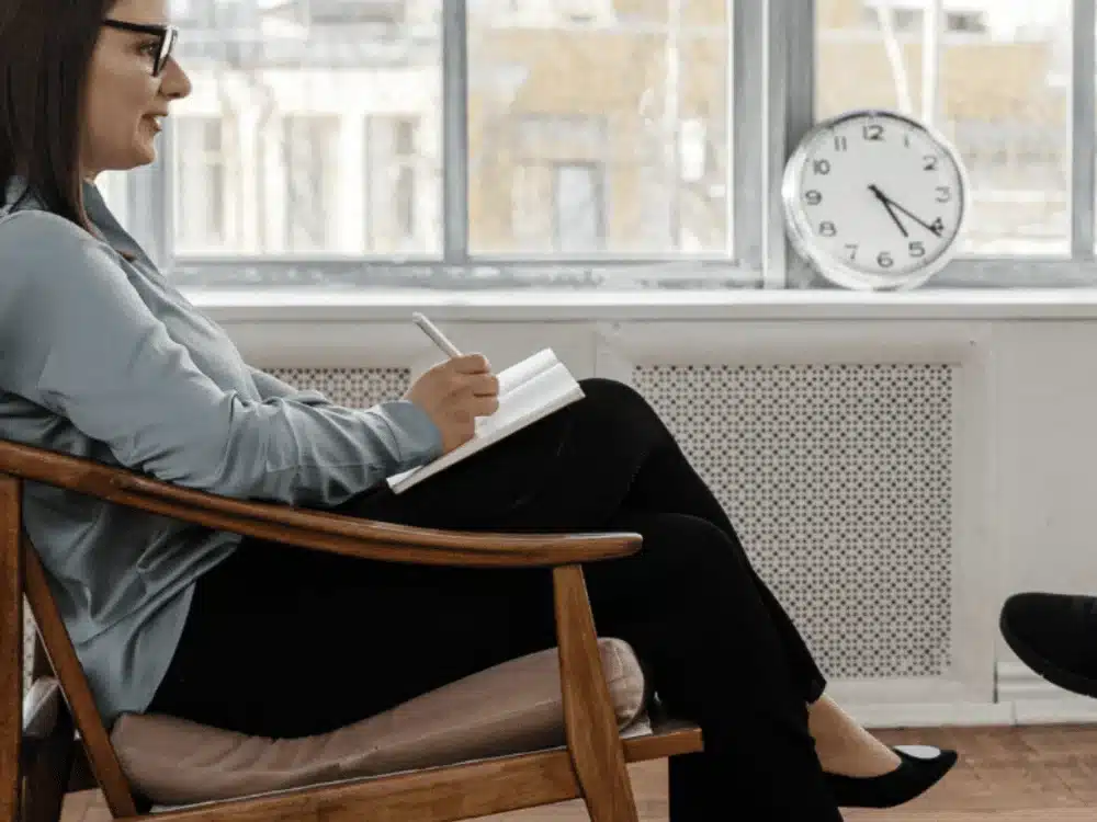 A person sits in a chair, writing in a notebook, with a clock on the wall and bright windows indicating a cozy indoor setting.