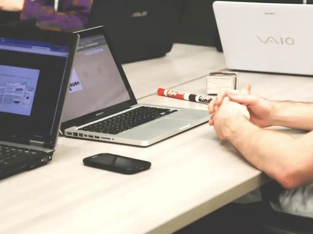 A person sits with folded hands beside laptops and a smartphone on a table, engaged in a collaborative work environment.