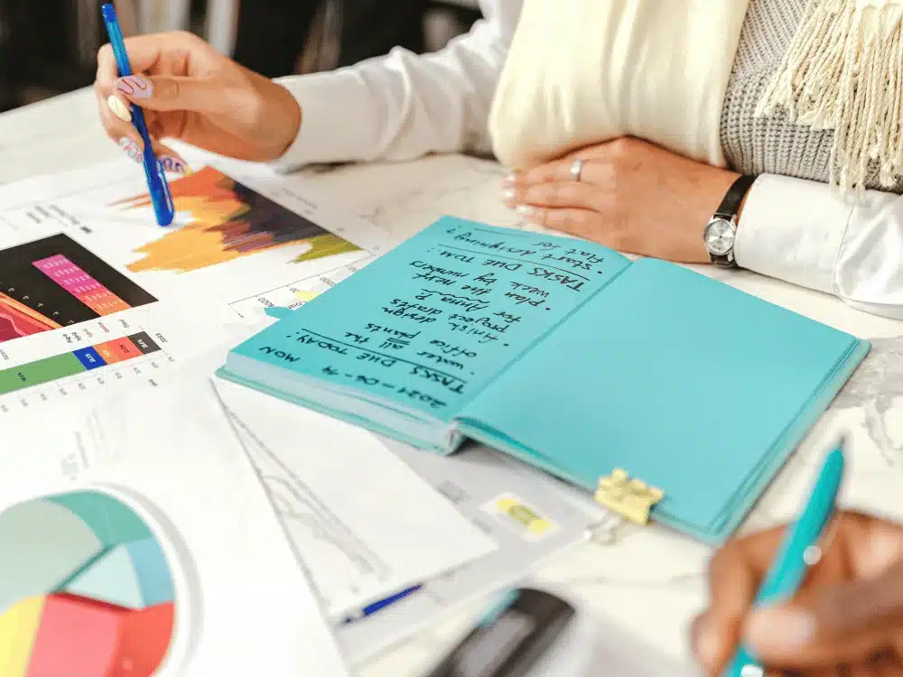 A person writes notes in a turquoise notebook while analyzing colorful charts and graphs on a table, surrounded by documents and pens.