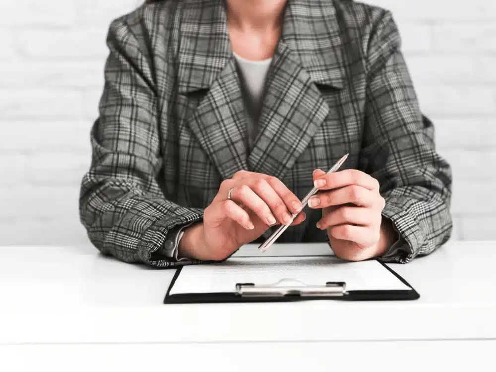 Detail of hands with pen and clipboard, with a plaid blazer in office setting.