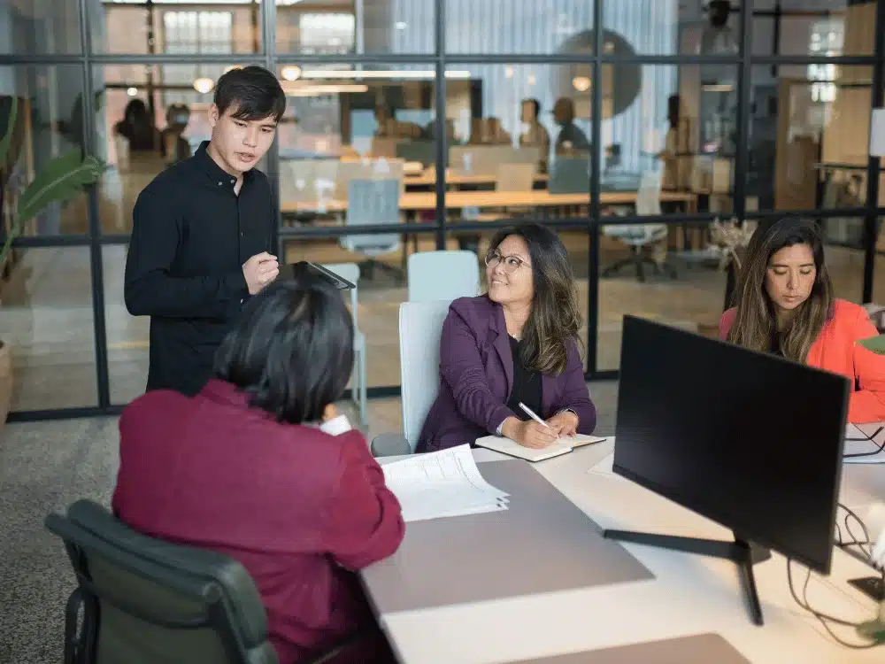 A business meeting with a man standing and talking to three seated colleagues.