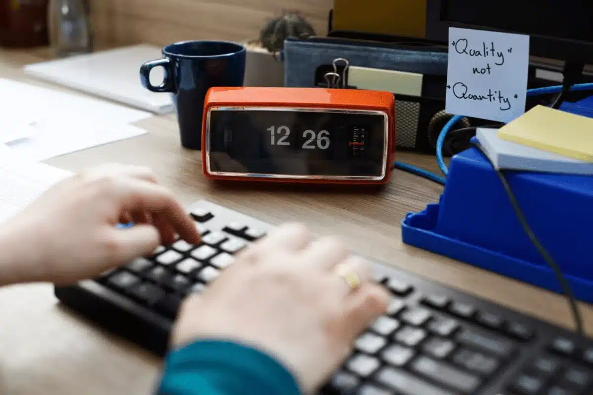 A hand types on a keyboard beside an orange clock displaying 12:26, next to a coffee mug and a note saying "Quality not Quantity."